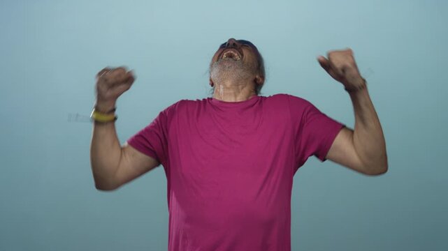 Man with hoary long hair, clenched fists and bare forearms shouting in a studio before a teal wall wearing magenta shirt; anger release.