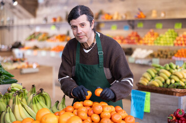 Obraz premium Adult man seller in apron puts fresh tangerines on display in vegetable shop