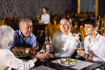Cheerful young couple spending time with aged friends in cozy restaurant. Girl and guy having fun while talking and enjoying light dinner with wine at table..