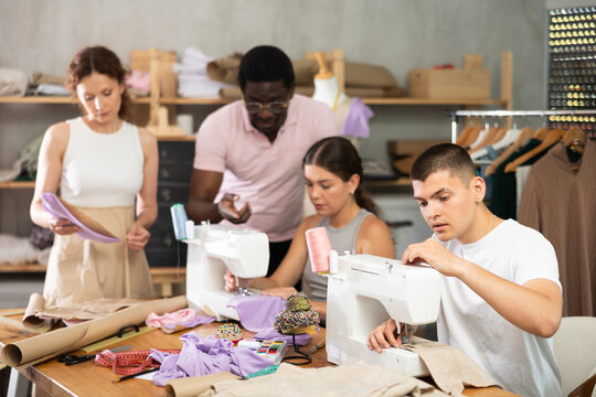Engaged young man enjoying hands-on learning during group sewing class for adults, stitching at machine in tailoring studio, while fellow learners sharing creative work and friendly conversation..