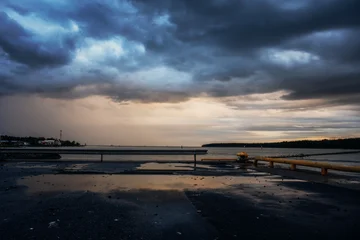Fototapete Gewitter Dramatic coastal landscape with dark storm clouds, sunset light and wet asphalt reflecting the sky. Moody seascape with harbor elements, calm water and a distant shoreline under heavy weather.  © Alexander