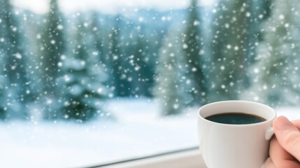 Hand holding a warm coffee mug by a window overlooking a snowy winter forest with falling snowflakes