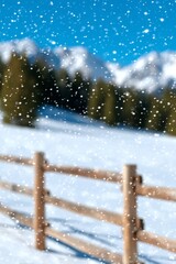 Snowy winter landscape with falling snowflakes, blurred mountains and evergreen trees in the background, and a wooden fence in the foreground