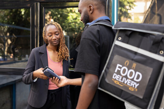 Delivery worker with backpack receives payment from woman for a meal order. The contactless nfc transaction with the smartphone reflects the modern online buying process for takeout services. - Powered by Adobe