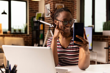 Smiling african american businesswoman seated at desk, having pleasant conversation on mobile device. Remote female employee taking break while using smartphone for video call in brick wall apartment.