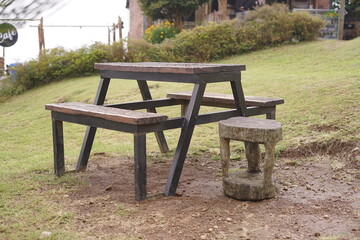 Rustic wooden picnic table with attached benches and a unique stone stool stands on a grassy patch, offering a tranquil outdoor seating area in a natural park