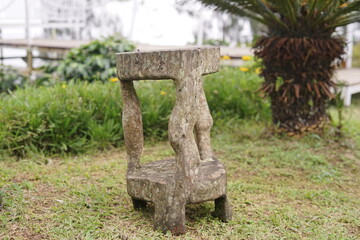 Weathered wooden stool with an organic, rustic design stands amidst green grass in a tranquil garden setting, offering a touch of natural artistry and unique outdoor decor