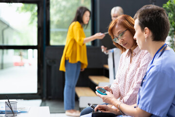 Nurse using glucometer to measure blood sugar during diabetes checkup in hospital lobby. Asian woman follows instructions while specialist prepares glucose meter and reviews readings.