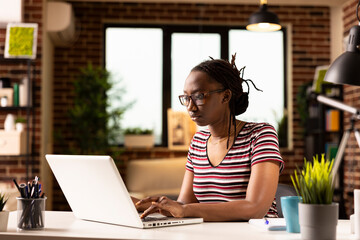 Black entrepreneur working remotely, managing tasks and reviewing financial data on laptop. African american woman with glasses, checking business reports and emails from home office.