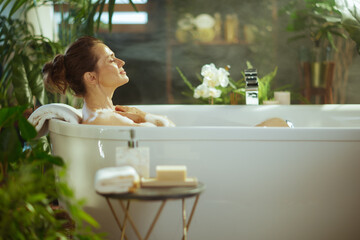 A serene woman with eyes closed and a peaceful smile relaxes in a white bubble bath, hands on her chest. The modern bathroom, with lush green plants & soft steam, creates a tranquil self-care oasis.