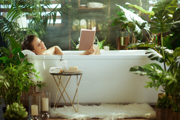 A happy woman finds peace reading in her white bathtub, surrounded by abundant green plants and soft candlelight. This serene image captures a moment of luxurious self-care and domestic tranquility.