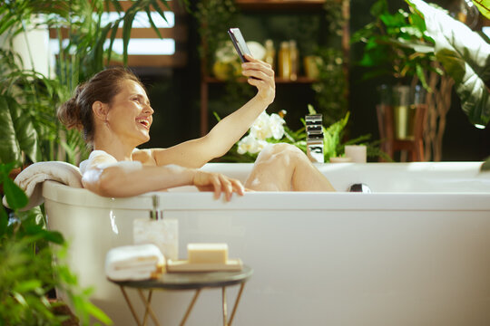 Smiling woman with a bun joyfully relaxes in a white bubble bath, holding smartphone for selfie. Lush green plants & bath products create serene, luxurious atmosphere for modern self-care moment.