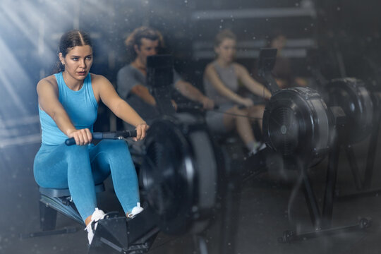 Sportive young girl training with rowing machine in sports hall during crossfit workout