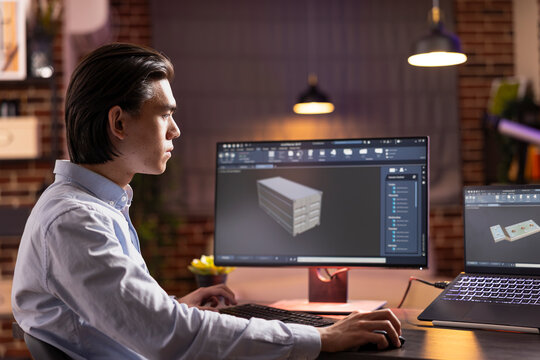 Asian male programmer manages tasks efficiently across multiple devices from apartment workspace. Focused young adult uses computer aided design software to develop and visualize industrial prototypes
