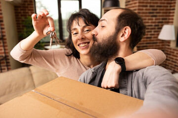 Happy caucasian woman dangling house keys and embracing boyfriend in new property. Bearded man kisses smiling wife on cheek and takes selfie, capturing joy during moving day in brick wall apartment.
