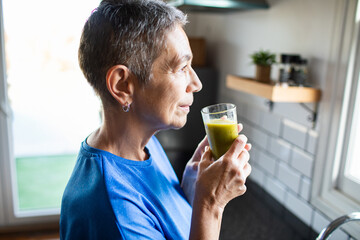 Senior woman calmly drinking green smoothie in home kitchen