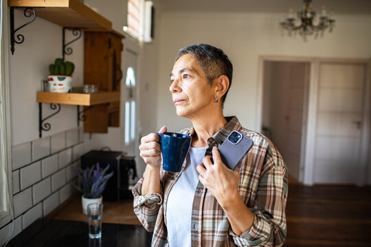 Senior woman holding coffee and smartphone, looking pensive in home kitchen