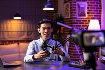 Creative asian man using digital camera to record podcast episode about wireless headphones. Male influencer filming professional social media video for web audiences and promotional online content.