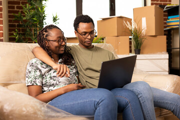 Cheerful couple sitting on sofa with laptop, planning new home after moving in. African American partners surrounded by unpacked boxes, browsing online for decorating inspiration in cozy living room.