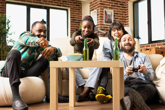 African american woman and caucasian man playing video game with wireless controllers as friends watch and point at screen. Diverse group of excited partners gaming together in modern living room. - Powered by Adobe