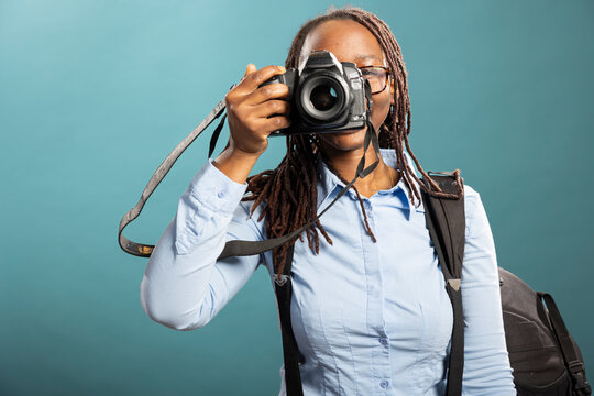 Professional photographer attentively working with her dslr in clean, blue background studio. African american woman wearing backpack, poses with her camera, capturing shots for client.
