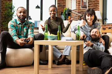 Bearded man and white woman team up with african american friends for online gaming. Multiethnic group reacting joyfully, holding controllers and enjoying fun video game in cozy apartment.