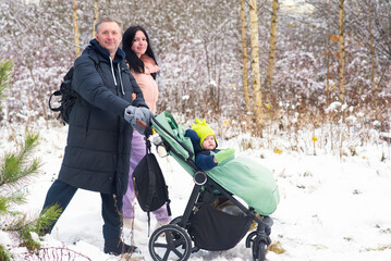 A family with a baby walks in a snowy park. A man and a woman push a stroller with a child. A father, mother, and baby boy in winter.