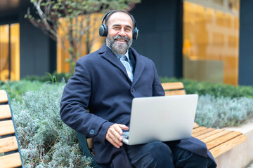 Mature man smiling using laptop and headphones outdoors