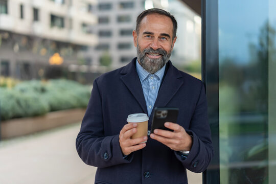 Smiling mature businessman holding coffee and smartphone outdoors