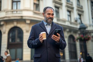 Mature man smiling using smartphone holding coffee cup in city