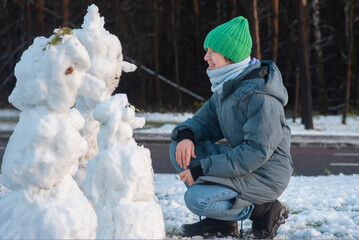 Teenage boy in grey jacket and green hat sitting facing small snowmen in winter, side view.