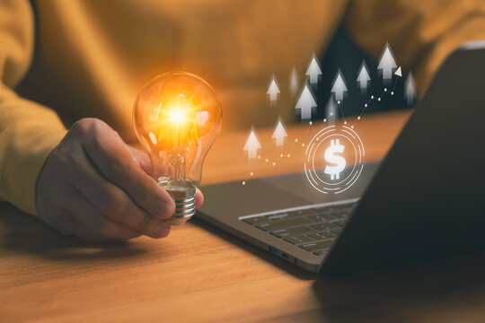 Innovation and business growth concept, person holding a light bulb beside laptop with rising arrow and dollar icon, symbolizing investing for the new generation, financial success, digital strategy.