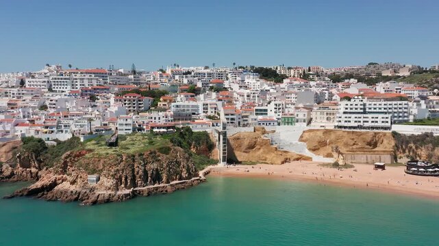 Coastal drone view of Albufeira with Praia dos Pescadores beach, dramatic cliffs, turquoise water, and vertical beach elevator connecting city to shoreline. Concept of urban beach access 