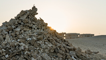 A tall cone-shaped mound of stones, coral debris, and seashells rises against the warm glow of the setting sun, creating a striking and atmospheric coastal scene.