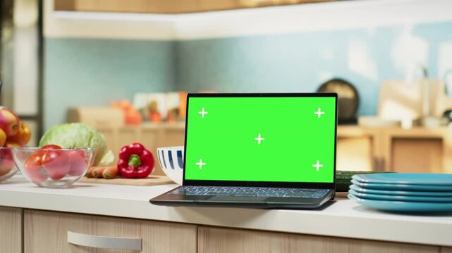 Green screen on laptop in modern home kitchen with cupboards and cookware on display. Countertop holds fresh organic produce and condiments, ideal background for cuisine or healthy eating.