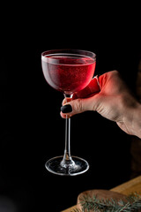A hand elegantly presents a bright red cocktail in a delicate glass, shimmering with bubbles. The dark backdrop enhances the drink's rich color, creating a striking contrast