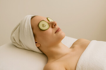 Horizontal spa portrait of a woman relaxing with cucumber slices placed over her eyes, wrapped in a soft towel and lying on a treatment bed, capturing a serene and refreshing self-care moment 