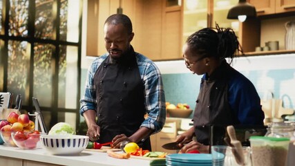 African American woman chopping red bell pepper on cutting board, preparing a fresh salad in the kitchen with her boyfriend. Girl preparing organic vegetables and ingredients for lunch. Camera B. - Powered by Adobe