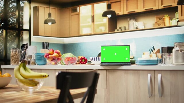 Green screen on laptop in modern home kitchen with cupboards and cookware on display. Countertop holds fresh organic produce and condiments, ideal background for cuisine or healthy eating.