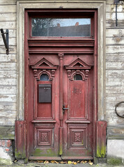 Close-up of an ornate, weathered wooden door with deep red paint, decorative carvings, and moss at the base, set in a rustic aged wall.