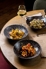 Three bowls of colorful pasta dishes are arranged on a wooden table beside a glass of white wine. The warm ambiance enhances the inviting culinary experience