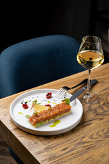 A beautifully arranged dish featuring a crispy item on a white plate, paired with a glass of white wine. The table is wooden, giving a warm and inviting atmosphere
