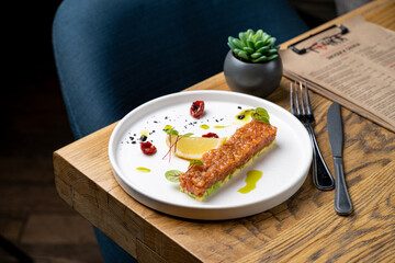 A beautifully arranged salmon dish sits on a white plate, garnished with herbs, lemon slices, and colorful reductions, against a warm restaurant backdrop