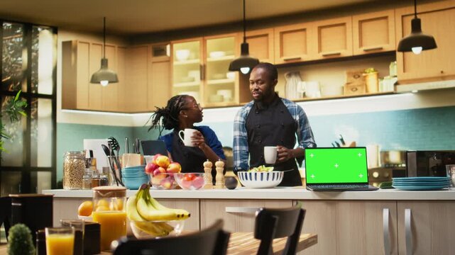 Green screen on laptop next to couple preparing vegetables for a salad meal, cutting ingredients and adding condiments in a bowl. Black people with aprons bonding in the kitchen. Camera A.