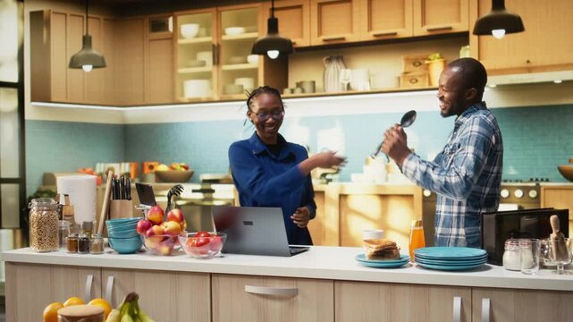 Overjoyed African American couple singing karaoke in the kitchen, showing positive energy and entertainment. People shows affectionate bond and creates a fun groovy atmosphere. Camera A.