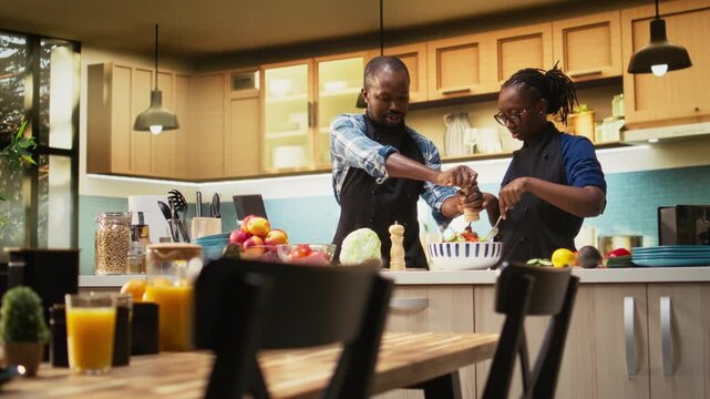 African american man adding pepper to the salad and woman stopping him, enough seasoning for healthy lunch meal in bowl. Young jolly couple preparing food together, wearing aprons. Camera A.