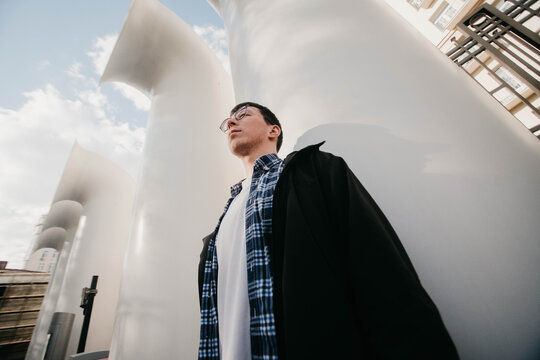 A young man showcases an air of confidence while leaning against sleek, modern architecture. The sunny afternoon highlights the unique design, creating an engaging urban scene