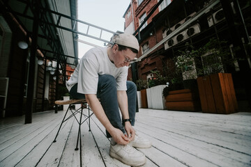 A young man sits on a chair in a modern outdoor space, carefully tying the laces of his sneakers. The scene features warm colors, wooden elements, and greenery, creating a relaxed atmosphere