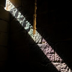Sunlight streaming through a narrow opening, casting a diagonal shadow on a textured stone wall with rough surface and natural color variations