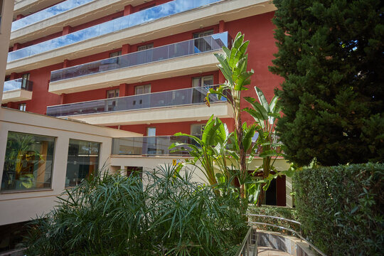 Modern hotel or apartment building with red facade and glass balconies, surrounded by tropical plants and greenery in Palma. architecture, vegetation - Powered by Adobe